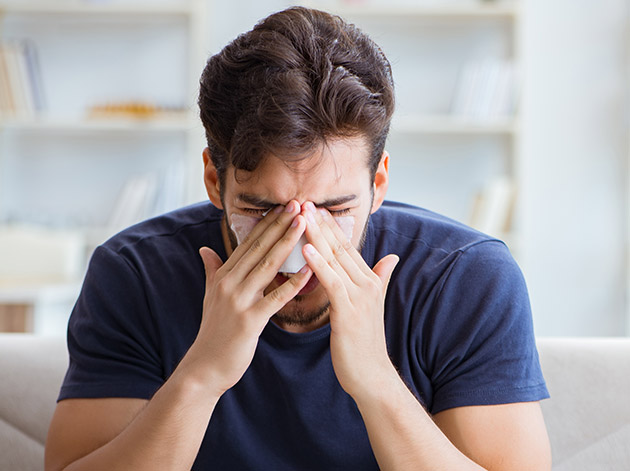 Man sitzt auf Couch, trägt dunkles T-Shirt, hält Hände vors Gesicht und wirkt nach einer Nasen-OP schmerzgeplagt.