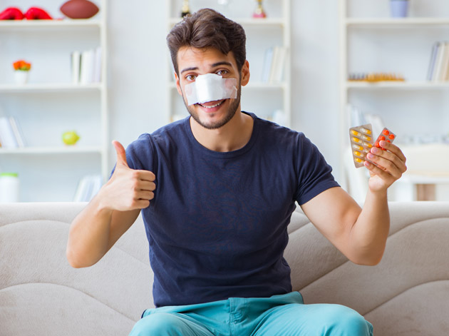 Man sitzt mit Nasenverband lächelnd auf Couch, zeigt Daumen hoch und hält Tabletten; Bücherregale im Hintergrund.