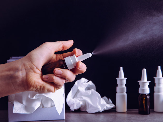 Hand sprüht Nasenspray neben Taschentüchern und Flaschen - Symbol für chronisch verstopfte Nase und Nasenspraysucht.