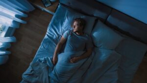Man liegt nachts wach im Bett unter blauer Decke, schaut zur Seite. Sanftes Licht von Fenster und Lampe.