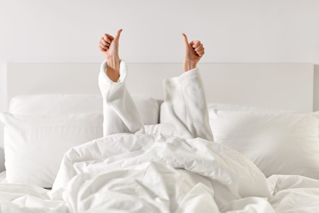 Person in weißem Bademantel unter weißer Decke gibt Daumen hoch nach einer Nasenoperation; friedliche, minimalistische Schlafzimmerszene.