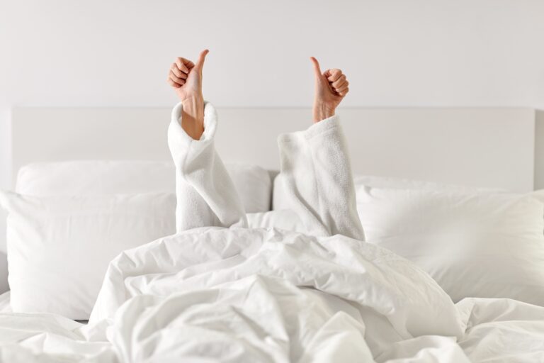 Person in weißem Bademantel unter weißer Decke gibt Daumen hoch nach einer Nasenoperation; friedliche, minimalistische Schlafzimmerszene.