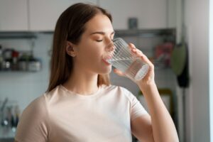 Frau mit langen braunen Haaren trinkt Wasser in der Küche, Augen geschlossen, entspannt, was auf Selbstfürsorge nach einer Nasenoperation hindeutet.